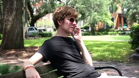 Young man sitting on bench outdoors in the park talking on the phone in slow motion. HD 720.