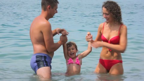 man and woman holds little girl splashing water by legs in sea 