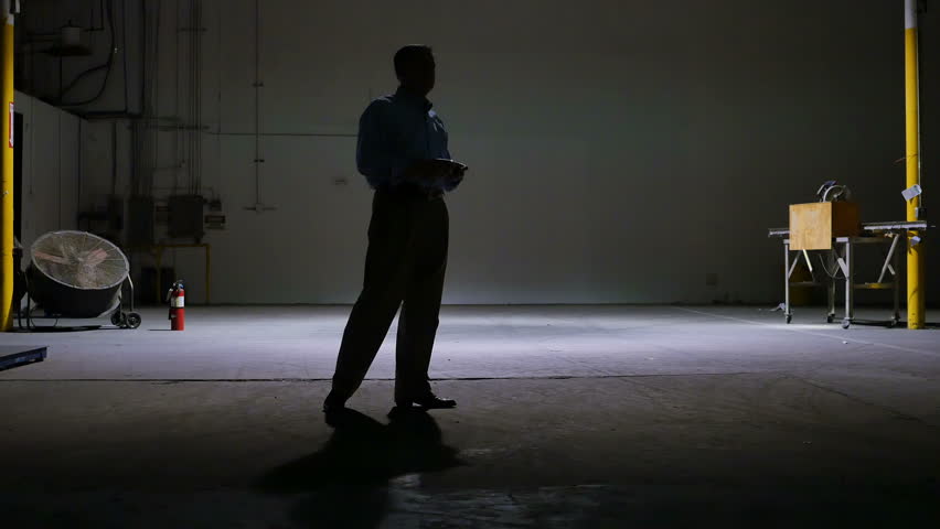 A man on a cell phone pacing in the lone light of a large dark empty warehouse.