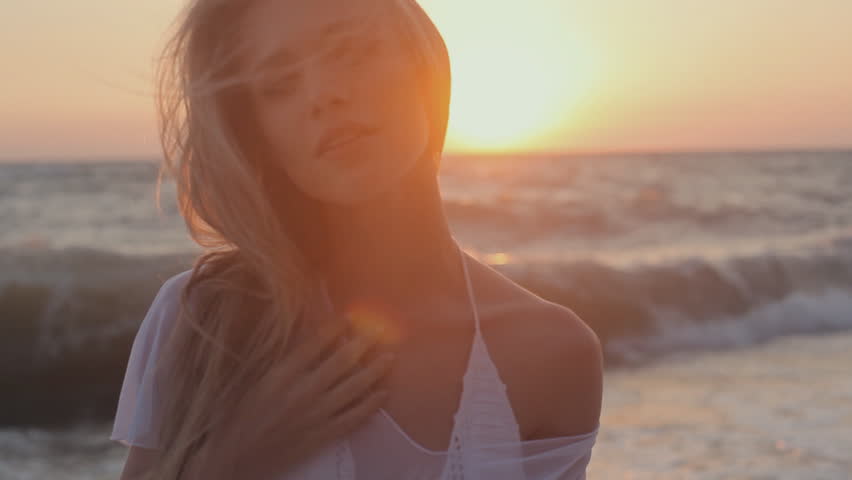 Seductive girl with long hair dressed in a transparent tunic posing at sunset on the beach