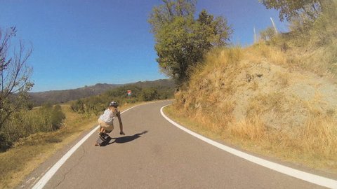 A longboarder performs tucking and sliding techniques while going downhill under a clear blue sky