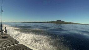 Wide angle view of Rangitoto Island, in the Hauraki Gulf, New Zealand, from a recreational power boat travelling through calm seas. Another motor boat and a stand-up paddle-boarder are nearby.