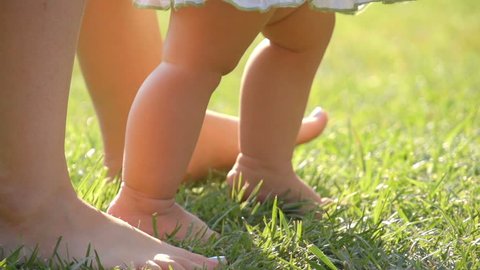 Little baby learns to walk. Mother is teaching her child to do the first steps on a green grass in summer. Close up on feet. Slow motion filmed at 250 fps. 