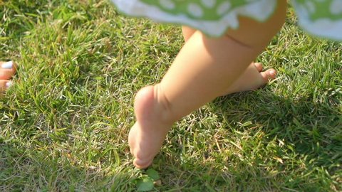 Little baby learns to walk. Mother is teaching her child to do the first steps on a green grass in summer. Close up on feet. Slow motion filmed at 250 fps. 