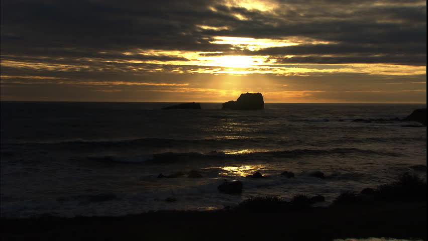 The setting sun silhouettes a large boulder in the sea