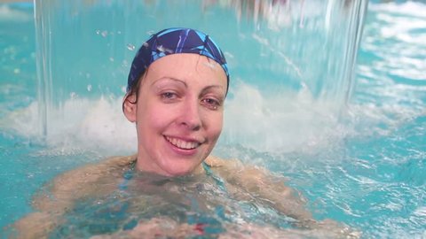 Smiling woman in swimming cap swims through waterfall in pool