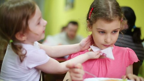 Little girl wipes face of other girl with white napkin in refectory