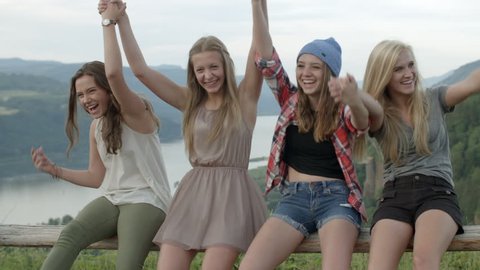 A Group Of 4 Teen Girls Sit On A Fence At A Beautiful Viewpoint, They Hold Hands And Raise Them In The Air  