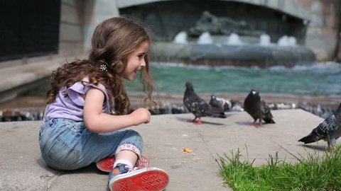Little curly girl sits and feeds pigeons near fountain at summer day