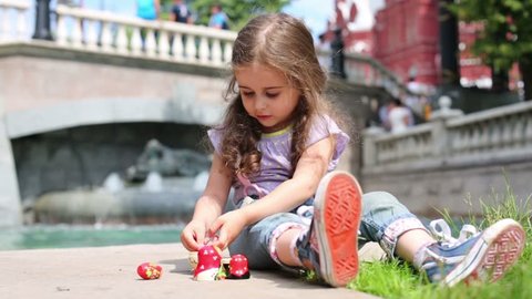 Little curly girl plays with matrioshka near fountain at summer day