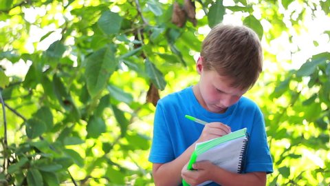 little boy sitting on bench near trees, boy writing in notebook
