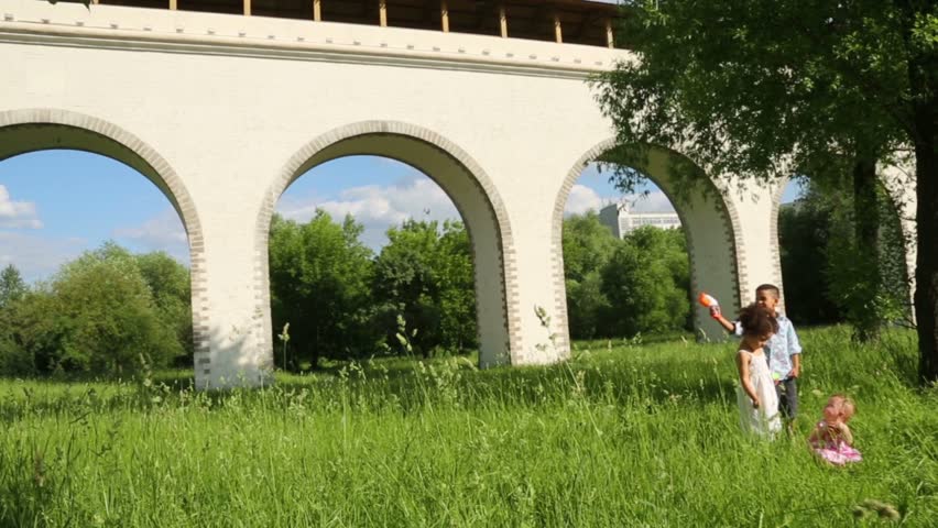 Boy with toy gun and girl wave hands, baby sits with apple on grass near aqueduct at summer