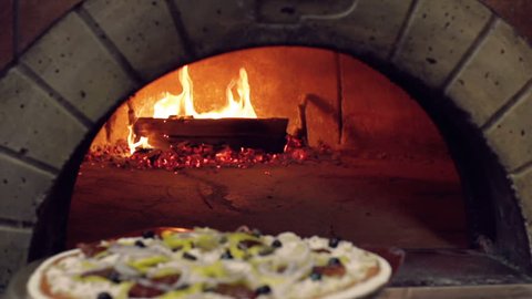 Tracking shot of unidentified cook placing pizza in wood-fired stove with pizza peel