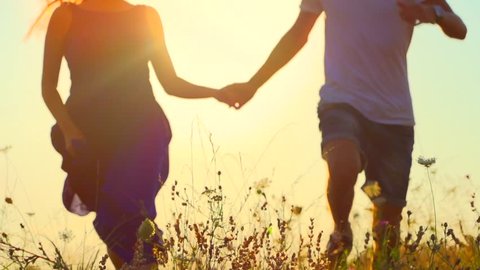 Happy couple having fun outdoors. Couple running away on the meadow. Countryside. Young Man and woman holding hands and running through a field with wild flowers. Sun flare. Slow motion video footage 
