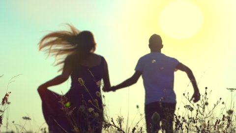 Happy couple having fun outdoors. Couple running away on the meadow. Countryside. Young Man and woman holding hands and running through a field with wild flowers. Sun flare. Slow motion. Love concept