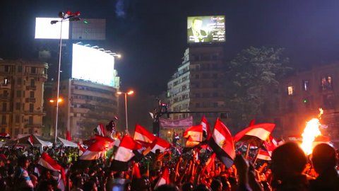View from the ground as protestors chant and wave flags at a large nighttime rally in Tahrir Square in Cairo, Egypt.