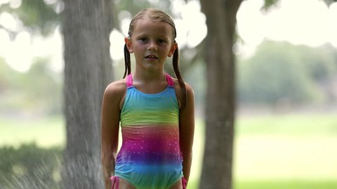 A sweet blond little girl in a colorful bathing suit stands in the water of a swirling water sprinkler and waits patiently.
