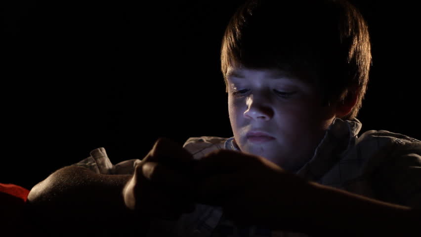 A young boy plays a handheld video game in the dark.  The light from the game