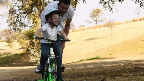 Steadicam shot of father teaching son how to ride his bike on a park pathway with safety helmet.