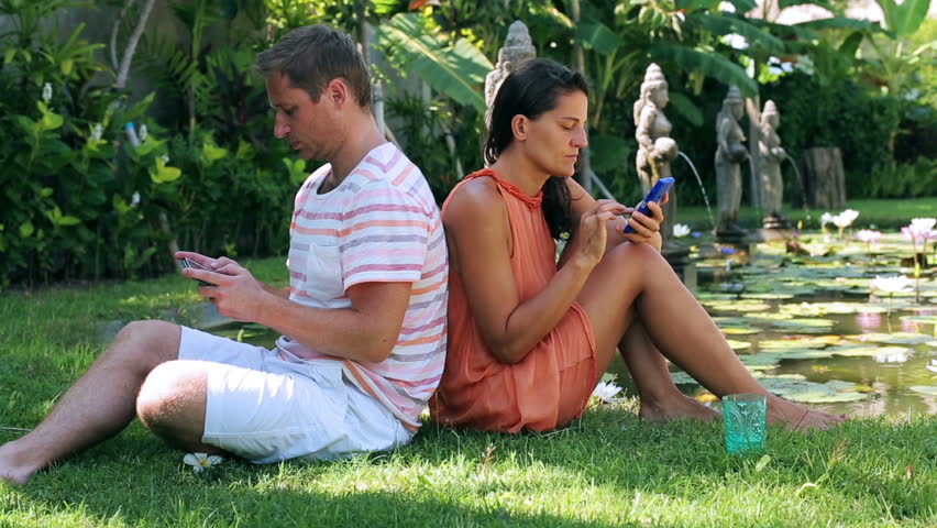 Couple sitting on grass in exotic garden and ussing cellphones

