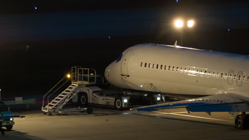 White Airliner Parked on an Overnight Lot Platform during the Evening with Ground Support Workers Carrying Out Tasks Around the Airplane.