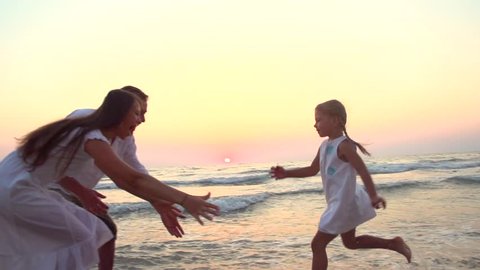Happy family having fun outdoors. Beautiful a little girl in a white dress running towards parents along coastline. Young happy family walking seashore Slow motion 240 fps HD 1080p. High speed camera