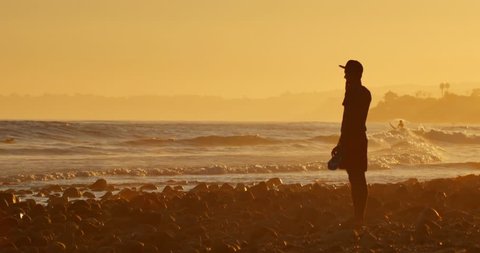 MALIBU, CALIFORNIA - August 29, 2014: Young man standing on ocean beach looking on waves of hurricane Marie at sunset. 4K UHD. Szerkesztői stockvideó