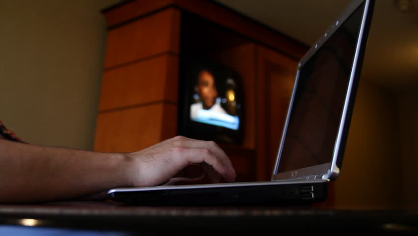 A man types on his computer in his hotel room as the television plays in the