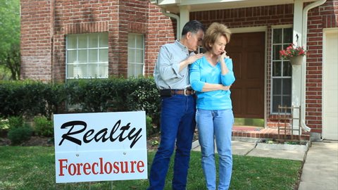 A distressed couple, standing behind a foreclosure sign in their front yard.