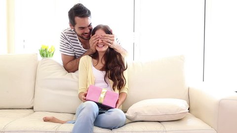 Handsome man surprising his girlfriend with a gift at home in the living room
