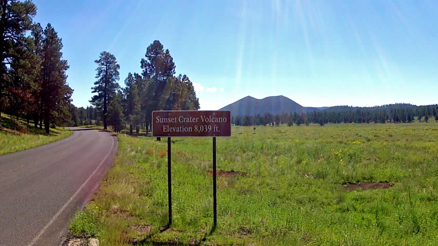 Shot of Sunset Crater Volcano sign next to the road. A wide vista provides a scenic view with inactive volcanic cone seen in the background.