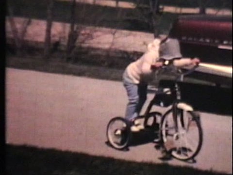 Three siblings ride tricycles and bikes in their drive way in front of some classic 60's cars on a crisp spring day.
