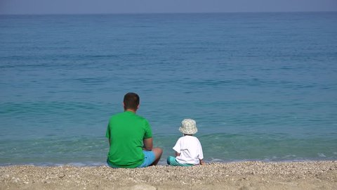 Child and parent sitting together on the beach at admiring turquoise sea Stock Video