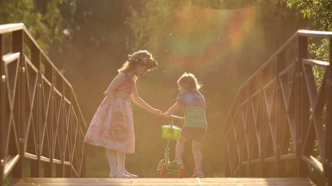 Two little pretty girls play with scooter on bridge at summer day