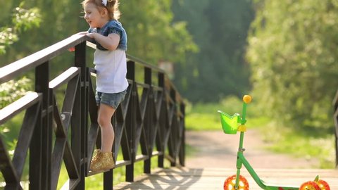 Little beautiful girl stands next to scooter on bridge and looks down at summer day