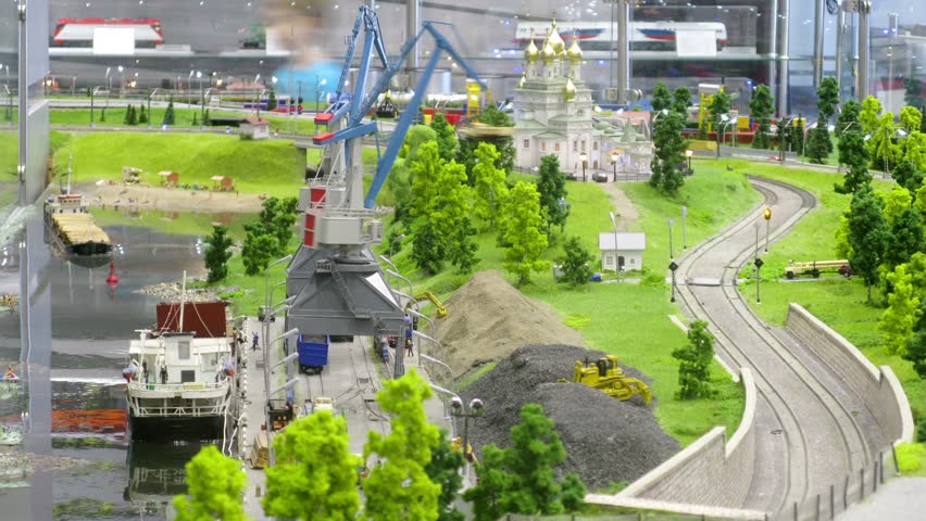 In modern toy city train bring tank-wagon on rail to storage station in port near transport ship and church. Time lapse
