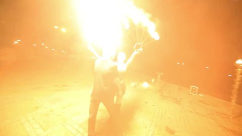 Guy fire breather spurts fireballs through burning fans in hands of girl.