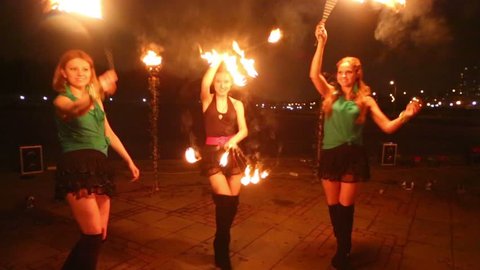 Three girls dance with burning fans at fire show in evening.