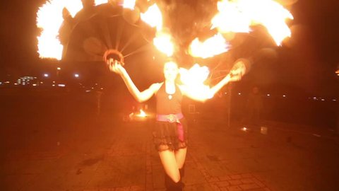 Girl dances with burning torches shaped as fans at fire show in evening, guy makes fireballs behind her.