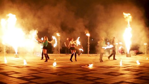 Group of firedancers dance with burning pois and fans, boy jumps making fireball.