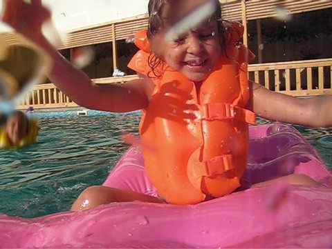 little girl sits on inflatable mattress in pool and splashes water to camera 