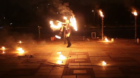 Girl moves with burning fans and boy rotates flaming pois at evening fire show.