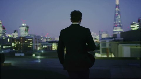 Successful businessman looks out at the view of the London city skyline at night