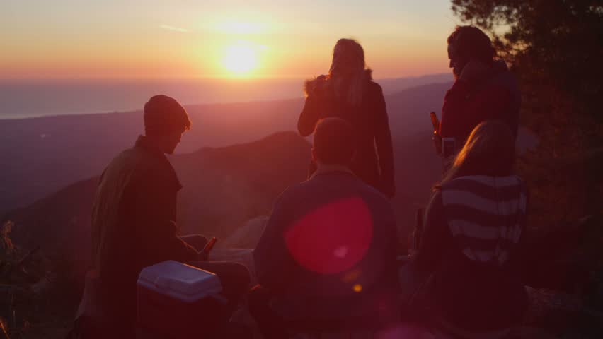 A group of friends drink beer at a campsite as the sun sets.