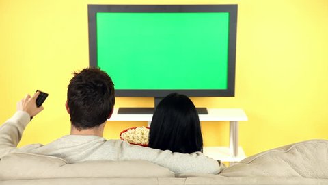 A young couple sitting on the sofa watching television and eating popcorn.  Screen is chroma-keyed so you can add your own video.