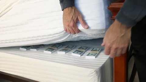 Man deposits a stack of bills to the money he is hiding under his mattress.