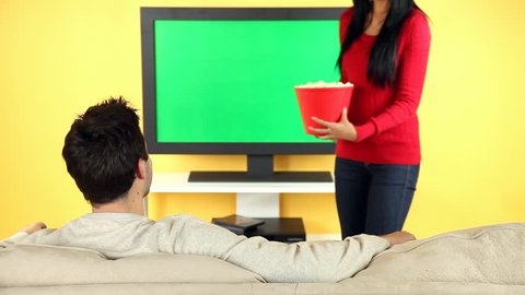 A young couple sitting on the sofa watching television and eating popcorn. TV screen is chroma keyed for adding your own movie or video.