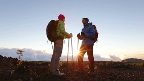 Hiking adventure healthy outdoors people standing talking. Couple enjoying sunset view above the clouds on trek. Video of young woman and man in nature wearing hiking backpacks and sticks.