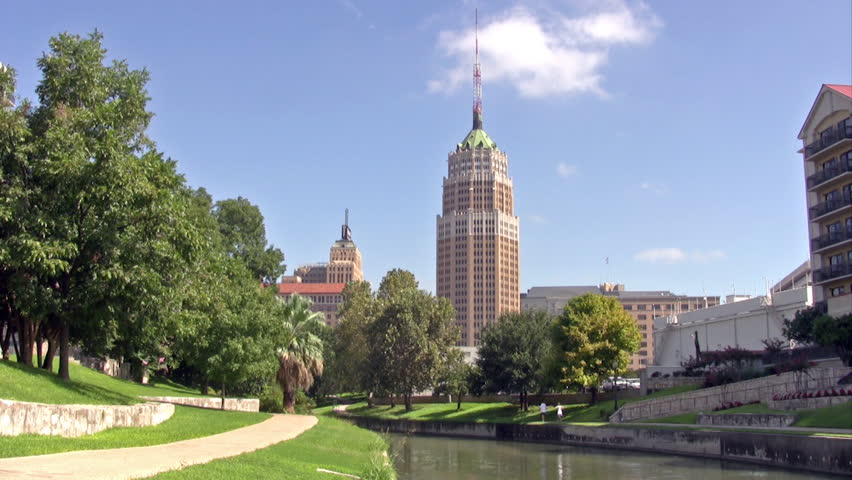 Video of the San Antonio Texas Riverwalk on the edge of the main city. Buildings, offices and hotel buildings. River flows through park with trees and walk. 