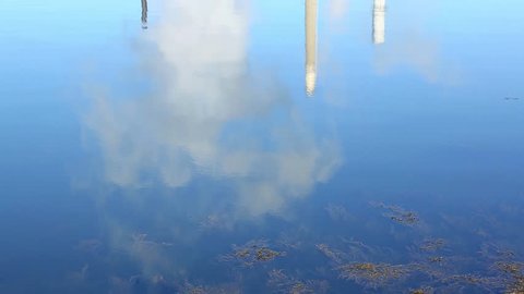 Video of reflected of steam rising from a factory and a bright blue sky with building and smokestacks onto the surface of sea water with seaweed floating in the foreground.